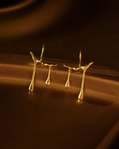 A pair of gold-plated sterling silver earrings designed in a minimalistic style with a geometric pattern, resembling a musical note, on a brown textured background.